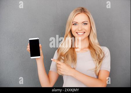 Copy space on her smart phone. Smiling young blond hair woman holding mobile phone and pointing at it while standing against grey background Stock Photo