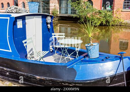 Manchester, Greater Manchester, UK. August, 24, 2024: A picturesque blue canal boat is docked on a serene canal, featuring a cozy seating area with a Stock Photo