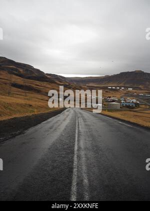 A wet road leads to a small town nestled among hills and mountains, under a gray sky, with power lines and utility poles seen along the route, capturi Stock Photo