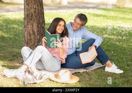 Couple enjoying leisurely afternoon reading books under tree in the park Stock Photo