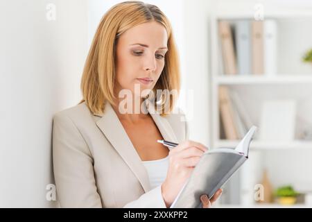 Everything must be in order. Confident mature businesswoman writing in note pad while leaning at the wall Stock Photo