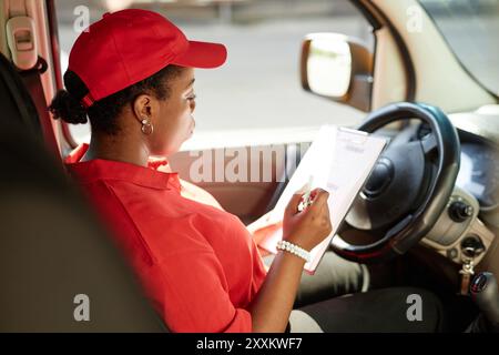 Black woman sitting in driver seat of delivery van, holding document with focused expression, getting ready for package delivery, sunlight illuminating her face Stock Photo