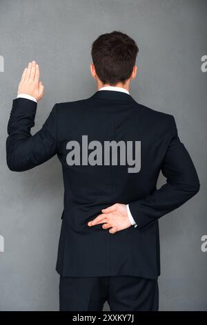 Telling lies. Rear view of young businessman keeping his fingers crossed and arm raised while standing against grey background Stock Photo