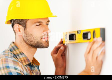 Checking this wall. Concentrated young repairman making measurements on the wall by level while standing indoors Stock Photo