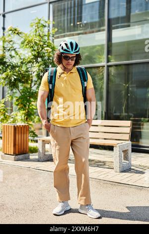 Young man with helmet and backpack is standing outside modern building and looking down with hands in pockets and sunlight Stock Photo