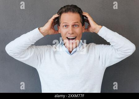 That is unbelievable! Surprised mature man touching head with hands and expressing positivity while standing against grey background Stock Photo