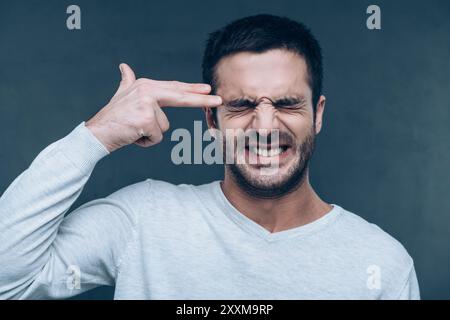 Bang! Frustrated young man gesturing finger gun near head and keeping eyes closed while standing against grey background Stock Photo