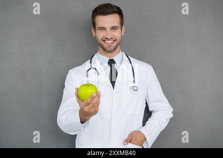 Vitamins are important for health. Cheerful young doctor in white uniform stretching out green apple and smiling while standing against grey backgroun Stock Photo