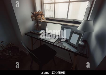A workspace with a dark modern aesthetic featuring a laptop, empty picture frames, a vase with flowers, and a lamp on a minimalist desk by a window, c Stock Photo