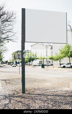 A large blank billboard mockup on a cobblestone sidewalk in a suburban or residential area. The background features dwelling buildings, parked cars on Stock Photo