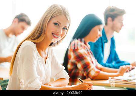 Working hard for good grades. Beautiful young woman writing and looking at camera while her classmates studying in the background Stock Photo