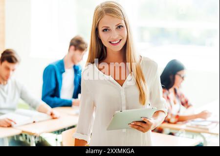 Tablet for making her study easier. Smiling young woman holding digital tablet and looking at camera while her classmates studying in the background Stock Photo
