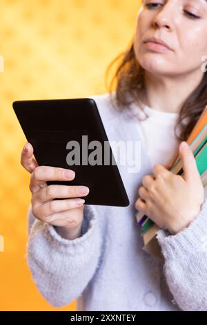 Woman carrying stack of books, reading online courses files for school exam on tablet, studio background. Student holding textbooks, doing research for university assessment on internet using device Stock Photo