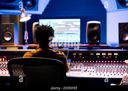 African american audio technician recording tracks from sound booth, processing tracks and working with digital software in control room. Expert pressing buttons and sliders for high quality. Stock Photo