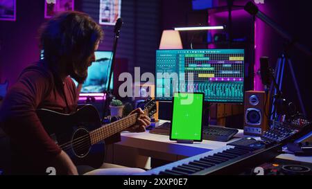 Male artist learning to play guitar on online lesson tutorial, practicing acoustic instrument and looking at phone with greenscreen display. Music producer doing rehearsal guitar play. Camera B. Stock Photo