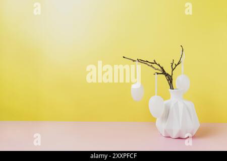 Happy Easter holiday, coming spring concept. Tree branches, flower twigs in white vase or jar with hanging eggs. Yellow and pink table background with Stock Photo