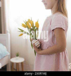 Easter concept. Small smiling girl in white fluffy bunny ears and pink dress holding yellow tulip flowers and colorful egg. Room interior. Stock Photo