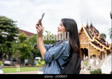 A beautiful, happy young Asian female tourist in a denim jacket and backpack is taking a photo of a temple with her smartphone, enjoying her visit in Stock Photo