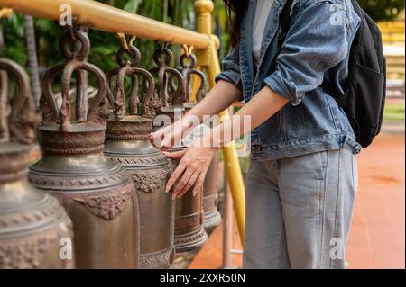 A close-up image of a female tourist in a denim jacket touching an ancient bell for luck in a Thai temple during her visit to Thailand. cultural trave Stock Photo