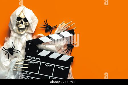 Scary evil skeleton smiling with movie clapperboard and spiders with copy space for text over orange background. Halloween movies concept Stock Photo