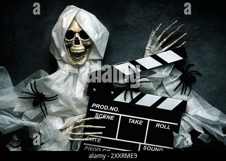 Scary evil skeleton smiling with movie clapperboard and spiders over dark background. Halloween movies concept Stock Photo