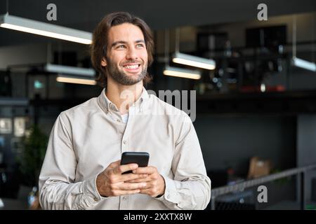 Young happy European business man entrepreneur holding phone standing in office. Stock Photo