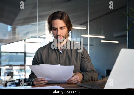 Busy young businessman executive holding documents using laptop at work. Stock Photo