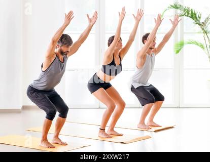 Group of people doing Utkatasana or Chair pose while practicing yoga in bright and spacious gym room Stock Photo