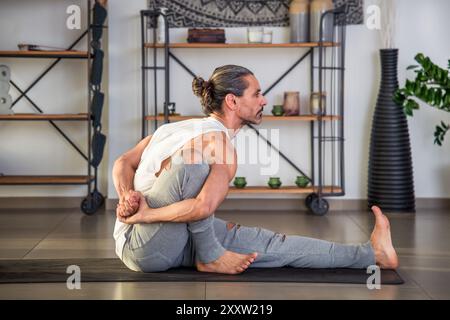 Side view of strong barefoot adult male athlete in sportswear looking away while sitting on mat and doing Marichyasana III yoga pose in hall during da Stock Photo