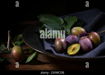 Freshly harvested plums with twig and leaves in a black bowl with napkin on a rustic wooden table, dark and moody food still life, copy space, selecte Stock Photo