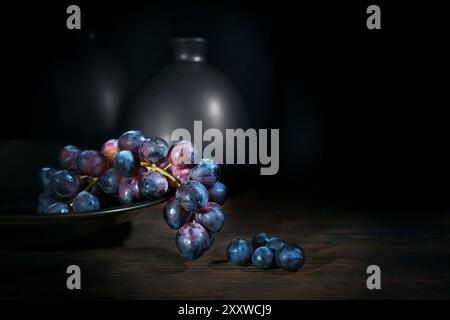 Purple blue grapes in a bowl on a rustic wooden table with vases against a black background, dark and moody still life with fruits, copy space, select Stock Photo