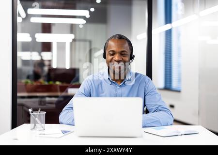 Confident businessman wearing headset engaged in online conversation on laptop. Smiling and communicating during virtual meeting in modern office setting. Concepts of technology, work, communication Stock Photo