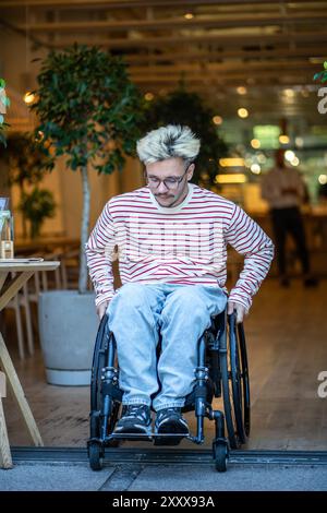 Serious focused man with disability in wheelchair drives out of cafe, trying to cross door threshold Stock Photo