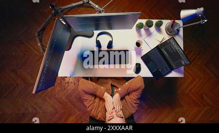 Top down view of fatigued man working overnight at home, rubbing eyes while taking break from coding, feeling sleepy. Tired IT admin exhausted from long screen time, pausing tasks to rest, aerial shot Stock Photo