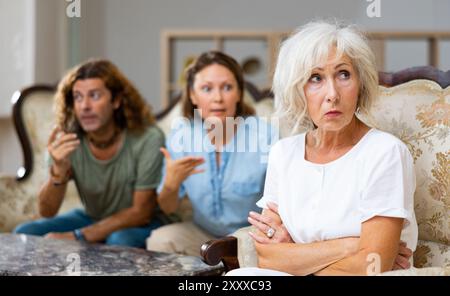 Angry mature female in armchair, keeping distance from the couple sitting on the couch at home Stock Photo