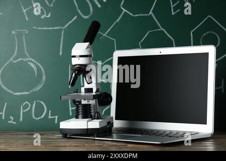 Modern microscope and laptop on wooden table near chalkboard with chemical formulas Stock Photo