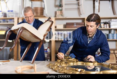 Mature male carpenter and his apprentice working on restoring vintage mirror and chair in workshop Stock Photo