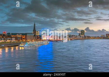 View towards Old Town of Düsseldorf with river cruise ship on river Rhine, Lambertus church, Schlossturm, and ferris wheel just after sunset Stock Photo