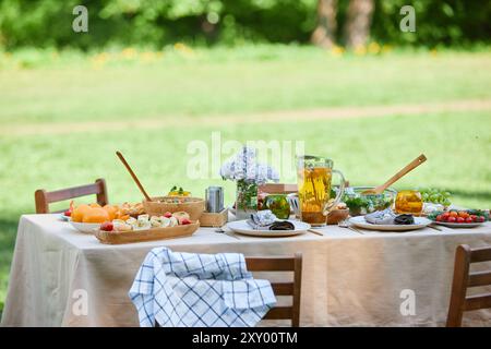 Outdoor picnic setting with diverse food and drinks displayed on linen-covered table in park under sunny weather. Wooden chairs surround table, creating a cozy meal area Stock Photo