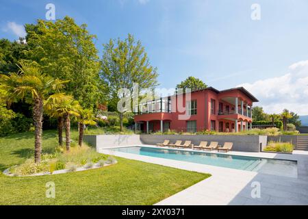 Red-walled villa in a big green garden with trees. There is a big swimming pool and sun loungers. Sunny summer day. Stock Photo