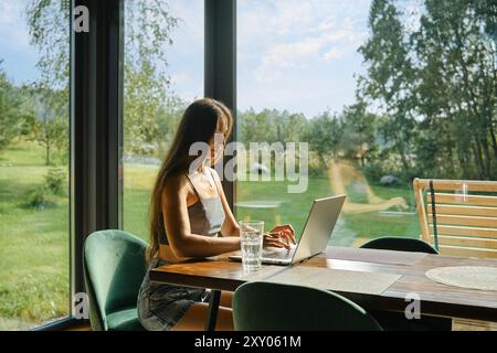 Woman with smooth long hair typing on laptop with lush green landscape outside. She is sitting in bright room with large windows of country house. Stock Photo