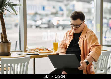 A man with Down syndrome smiles while using a laptop at a cafe table with a slice of pizza. Stock Photo