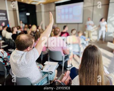 Audience member raising hand during conference session, indicating engagement and interaction in professional setting. Presentation takes place in modern conference room with attentive participants. Stock Photo