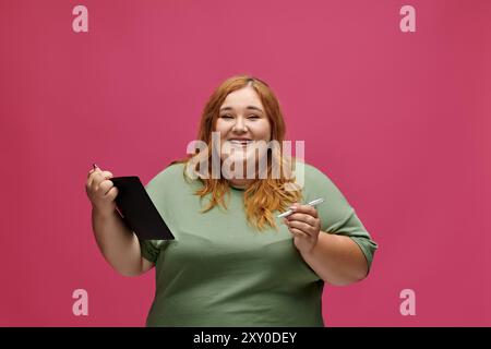 A plus size woman smiles while holding a notebook and pen in front of a pink background. Stock Photo
