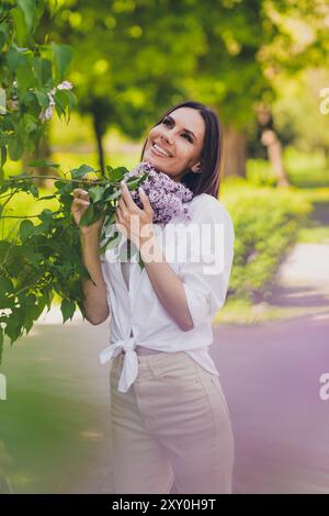 Vertical photo of young cheerful woman posing dreamy smell lilac flowers wear white outfit walk park sunny summer weather outside Stock Photo