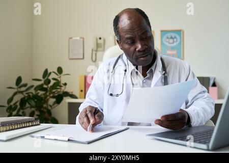 African American doctor sitting at his desk, reviewing patient documents carefully with stethoscope around neck. Workspace includes laptop, plants, and office accessories Stock Photo