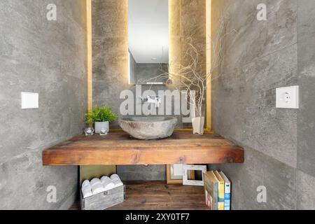 A chic, modern bathroom featuring a rustic wooden countertop, stone sink, and sleek concrete walls illuminated by warm, ambient lighting Stock Photo