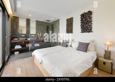 A tastefully decorated modern bedroom featuring a glass wall that integrates the bathroom, enhancing the sense of space Wood and stone elements contri Stock Photo