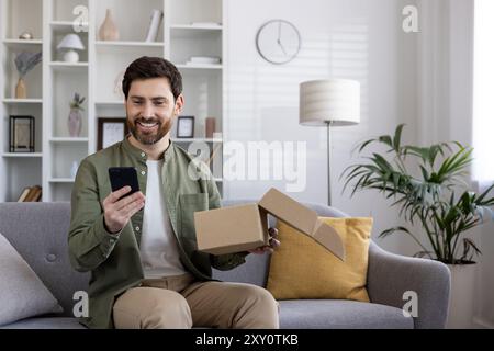 Smiling man checks online delivery with phone on couch. Holds cardboard box while engaging with smartphone technology. Bright living room setup, casual and relaxed atmosphere, natural lighting. Stock Photo