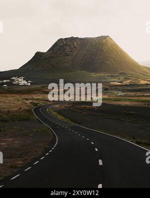 A scenic view of the Cuervo volcano and a winding road with a lone car driving towards it, nestled in the dramatic landscape of Lanzarote, Canary Isla Stock Photo